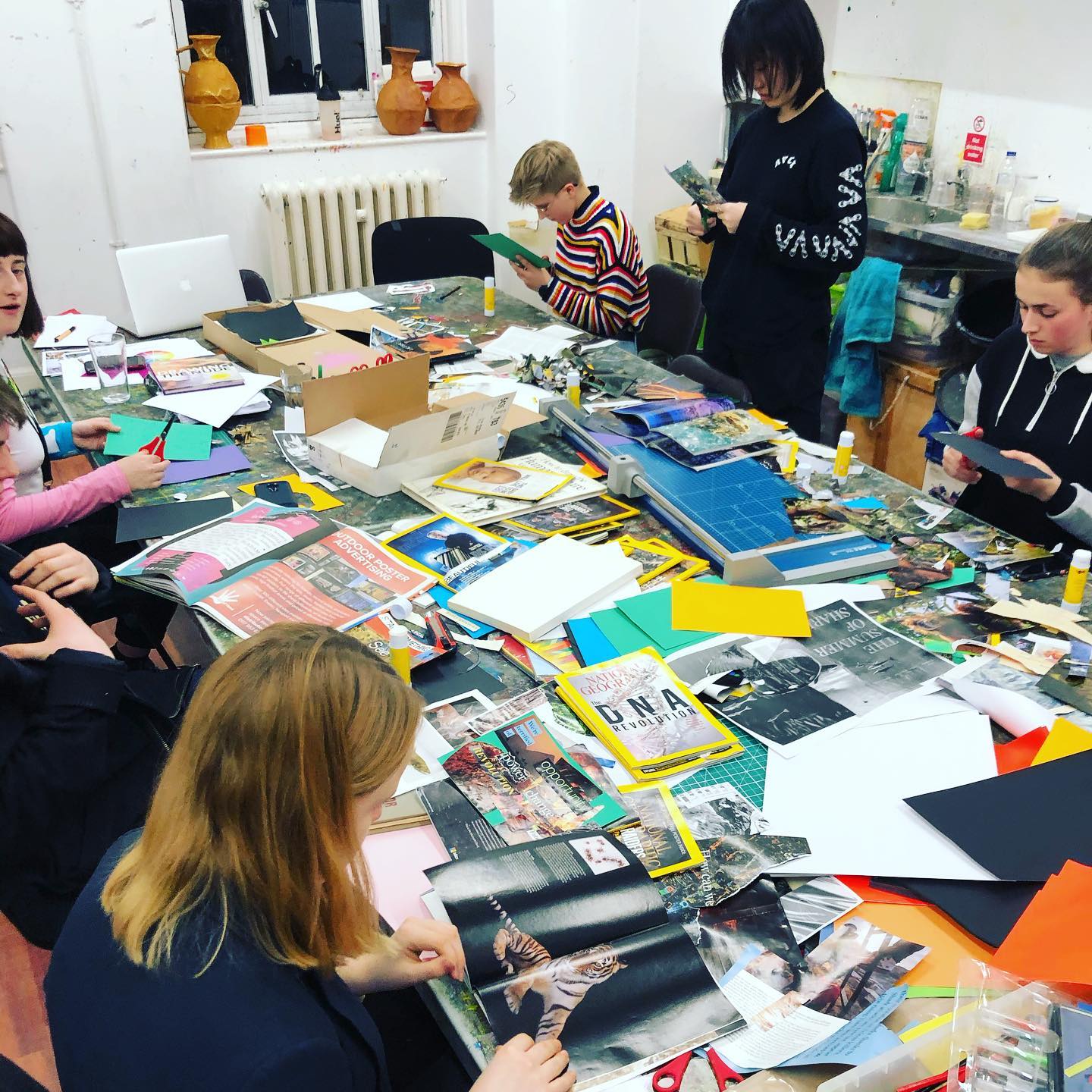 88433637_3532689573473060_1315623636102545408_o A group of young people are cutting up paper on a messy art table.