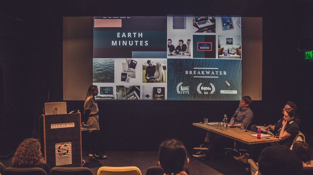 A woman stands in the cinema to pitches a film idea to three people sat at a table