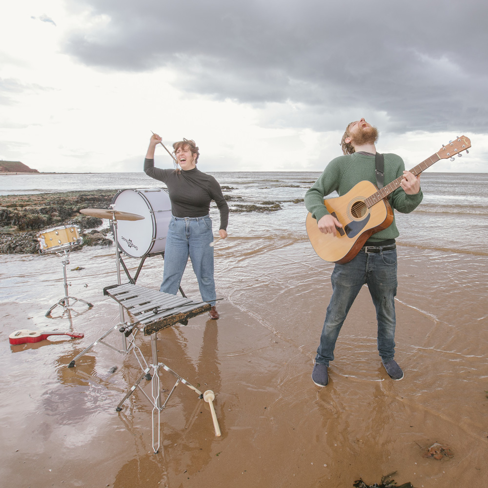 An image of two performers on a beach. A female performer is hitting a drum. A male performer is playing guitar and shouting,