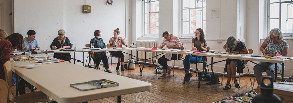 A group of artists sit at a U shape desk and work on some artwork.