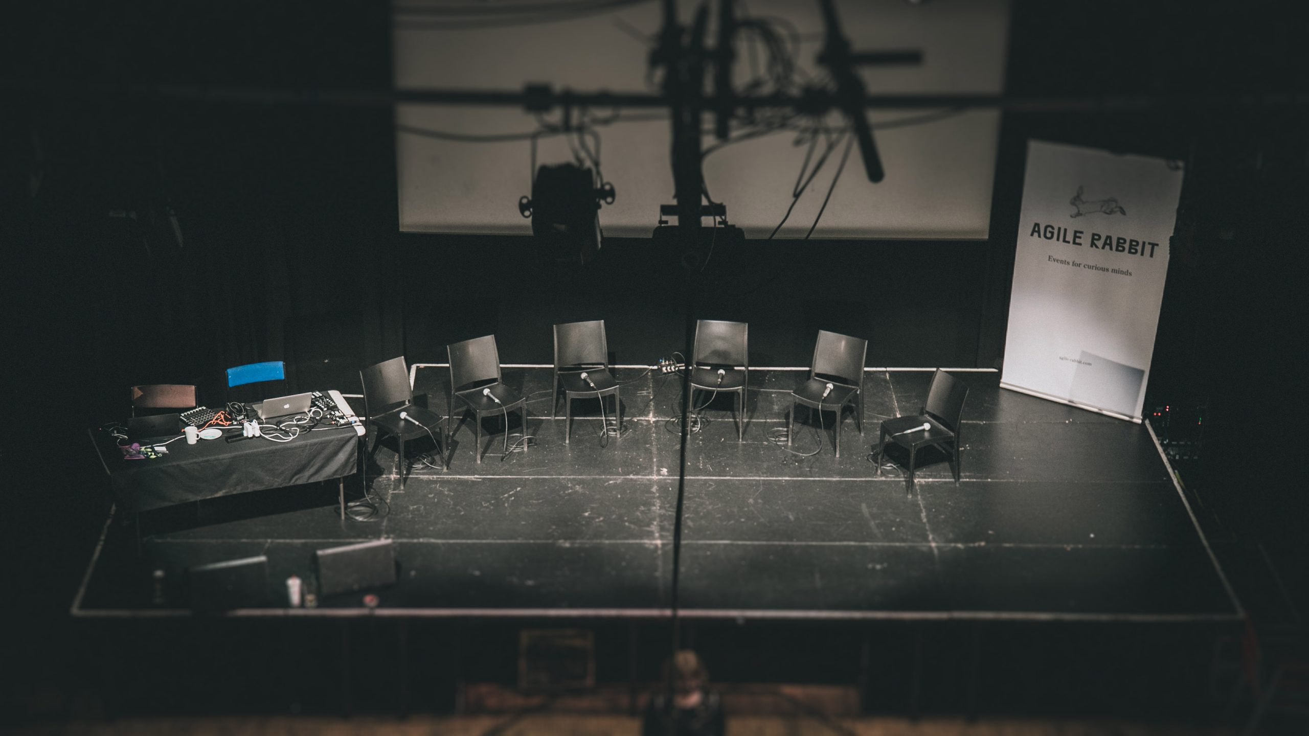 A spotlight shines on an stage that is empty apart from 6 chairs and a desk