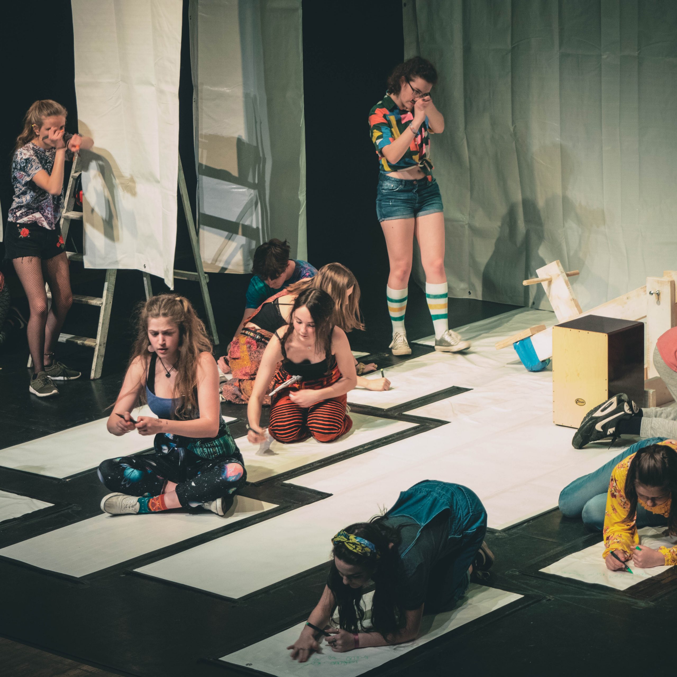 Photo of several young people taking part in a theatre show, many of them are drawing on large pieces of paper