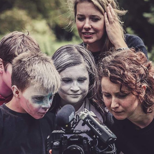 A small group of children gathered around a video camera watching back something they have filmed