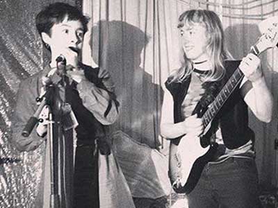 A photograph of someone playing a guitar while someone else speaks into a microphone.