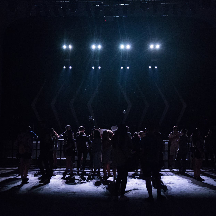 Dancers stand on a dark stage, back lit with spotlights