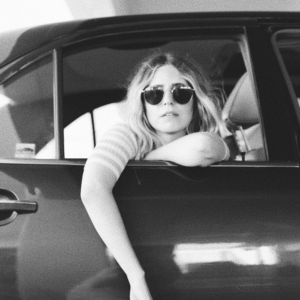 A black and white image of singer Joni facing out of a car window wearing sunglasses