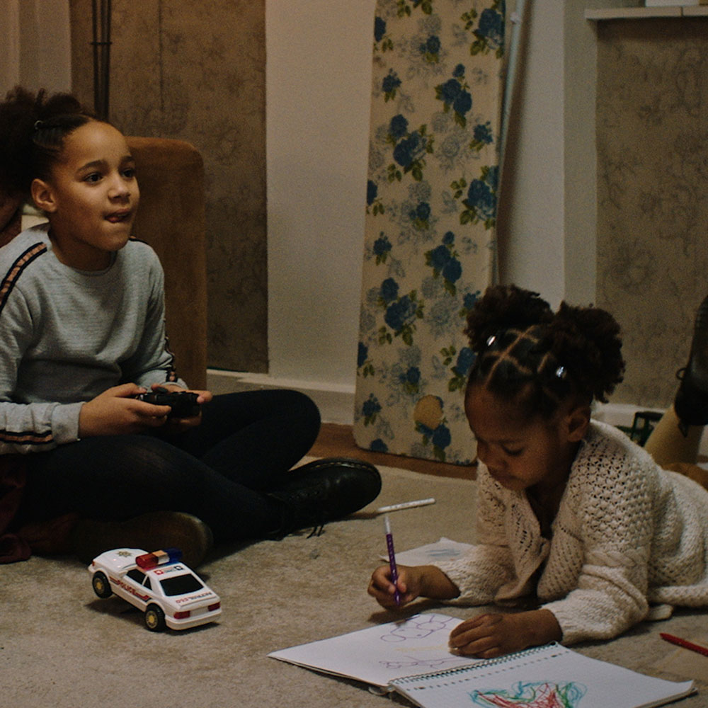 Two girls sit on the floor. one plays a game and one draws in a sketch book