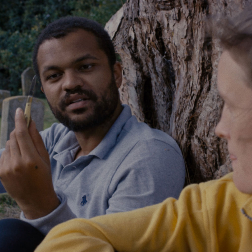 A man in a blue shirt holds a vial up as he looks at a woman in a yellow jumper