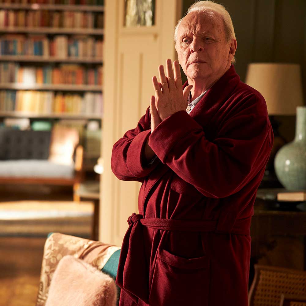 Anthony Hopkins stands in a living room in a red dressing gown. Behind him are large bookshelves filled with books.