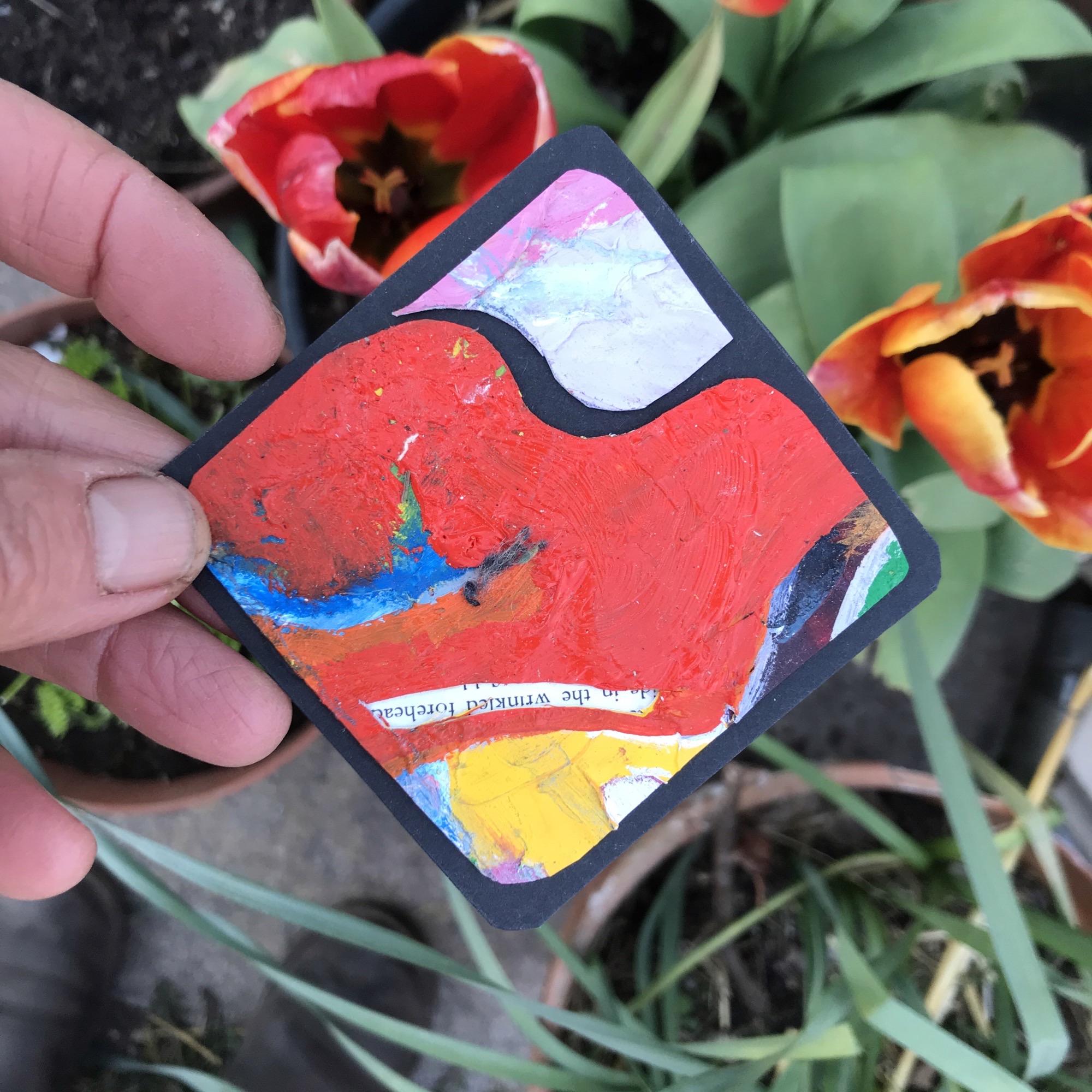 A small square mat, painted into three main sections. The middle section is painted mostly red with a splash of blue. The top section is silver with a slither of pink and the bottom section is a mixture of yellow, blue, pink, silver, red, orange and green. This mat is being handheld above two bright tulips outside.