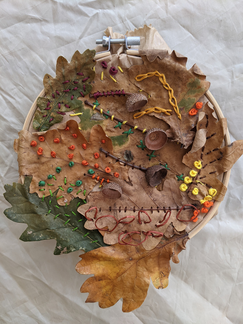 A group of green, orange and brown leaves in a circular basket taken from a birds-eye-view. The leaves' natural patterns are accentuated by women thread, small balls of thread and acorns.
