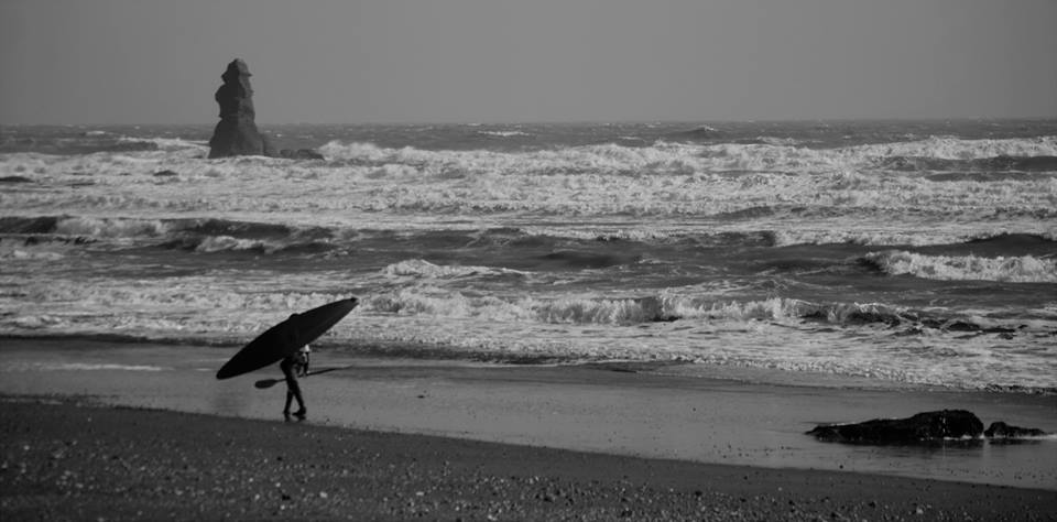 A person who's upper body and head is obscured by a paddle board walks across a beach holding a paddle in the opposite hand. The picture is black and white and the person is on the left of the image walking right.