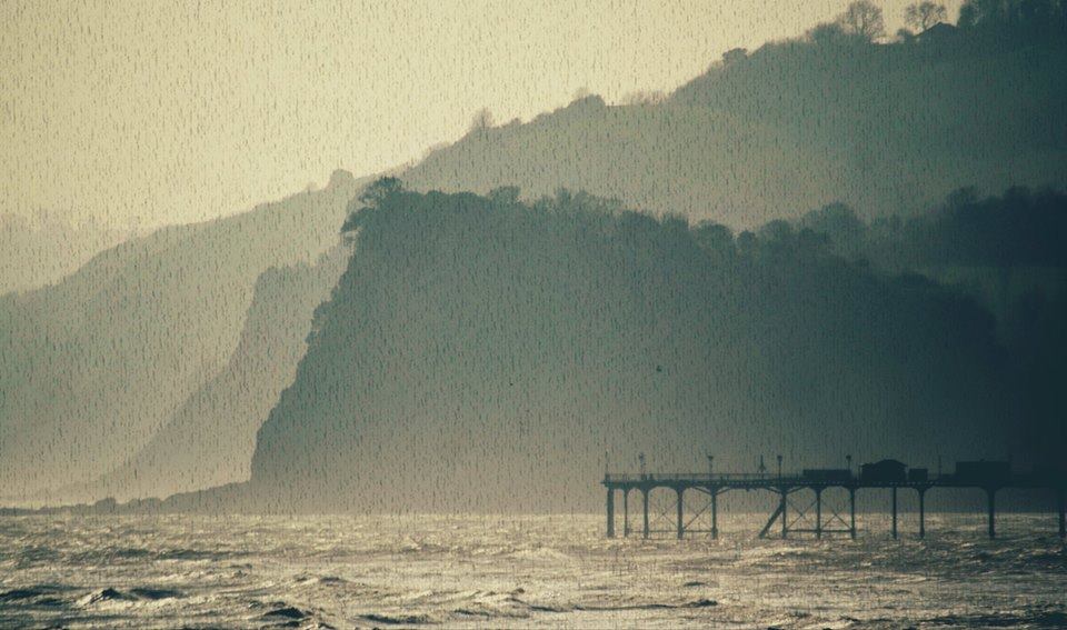 A grainy ektachrome image presents large silhouettes of cliffs. Underneath them there is an edge of a pier standing in the sea.