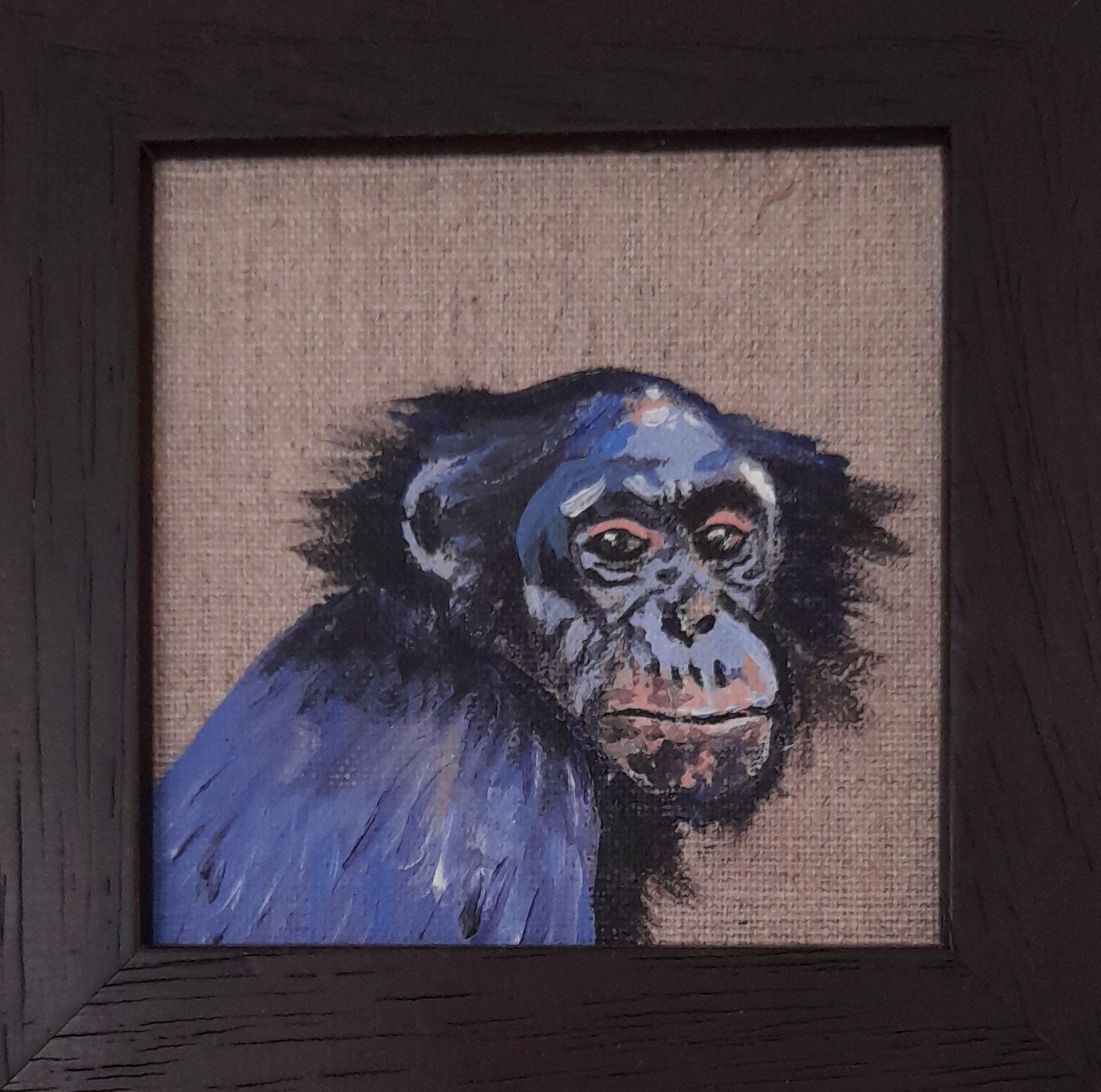 A blue baboon is painted onto a brown canvas and looks out of a black frame.