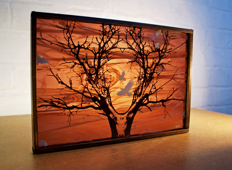 A golden-framed, landscape canvas standing on a wooden table. On the canvas, there is a transparent deer's head with antlers growing into branches of a tree. The background is made up of orange swirls and there are a few grey birds flying between and around the branches.