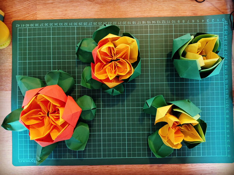 Four card-crafted flowers are photographed from a birds-eye-view, sitting on a measuring mat. The sizes of the flowers decrease from left to right as the colour grade of each flower gradually changes from orange to yellow. All flowers have green sepals.