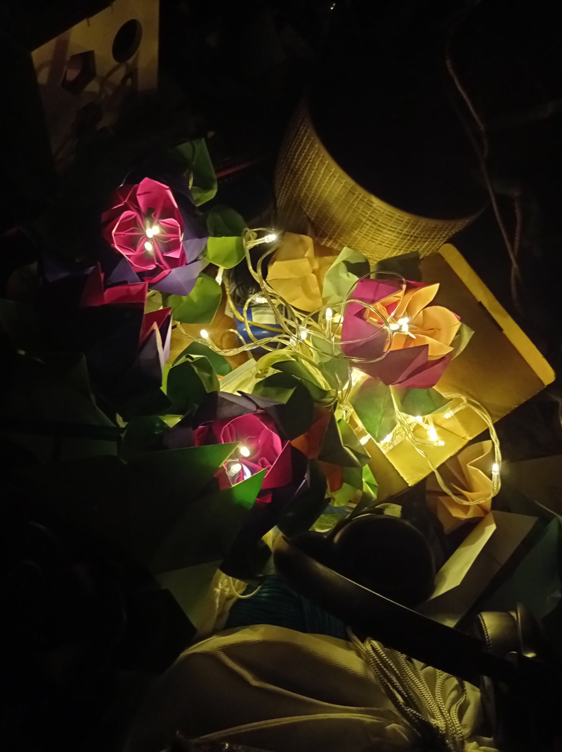 Coiled fairy lights are lit and wrapped around three card-crafted flowers in the middle of a dark room.