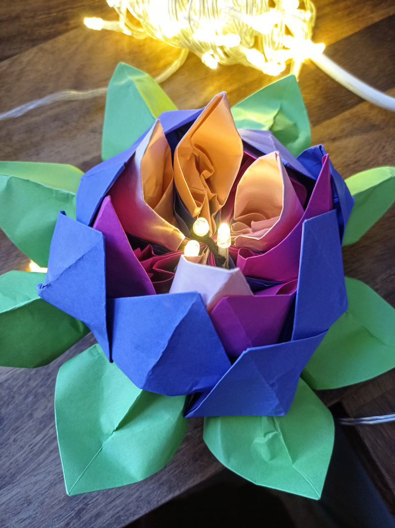 A close up picture of an iris-coloured, card-crafted flower with green petals. Inside are three lit-up fairy lights to resemble its anthers.