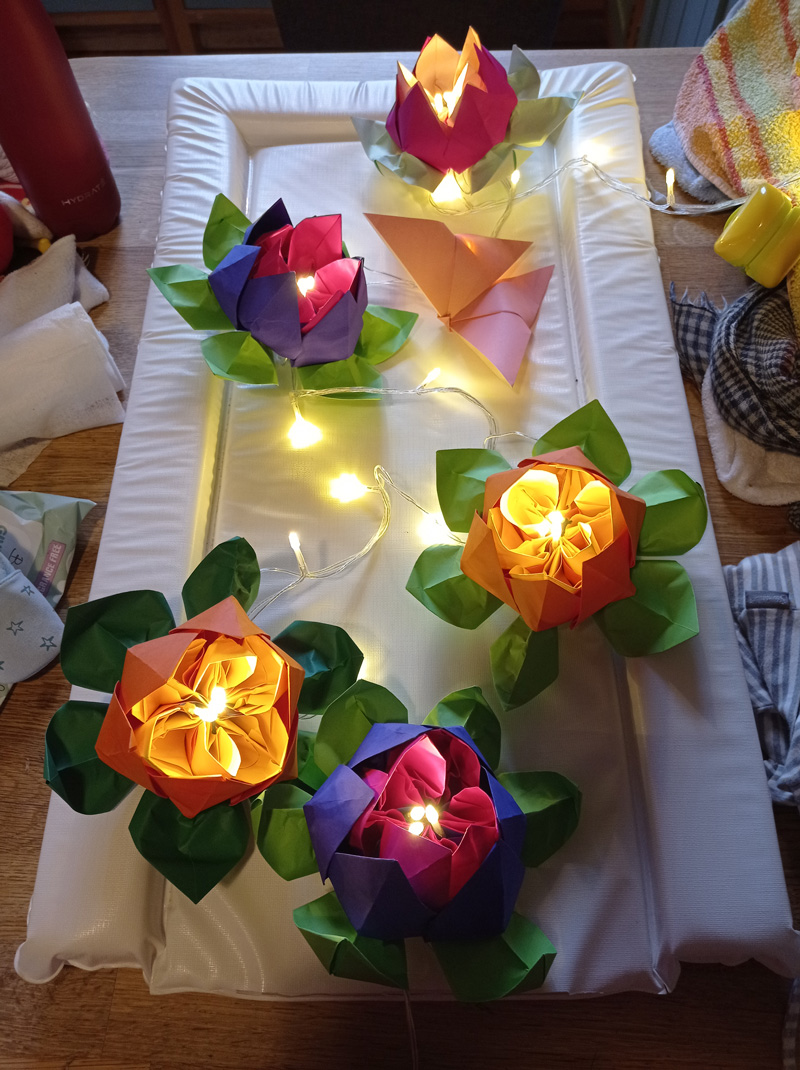 A picture taken above of five card-crafted flowers with fairy-lit centres. From front to back, the first and fourth flowers are purple-coloured, the second and third flowers are salmon-pink and the last flower is a warm violet colour.