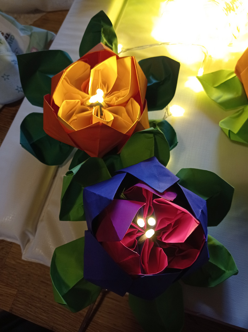 Two card-crafted flowers have three, lit fair lights in their centre. The top-left flower is orange and the bottom-right is purple. Both have dark green sepals.