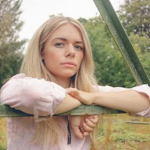 Chloe Foy leaning on a fence looking t the camera in a pink top