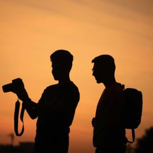 The silhouettes of 2 men holding cameras against the setting sun.