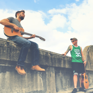 A man with a guitar sits on a wall. Another man in a green top stand beside him