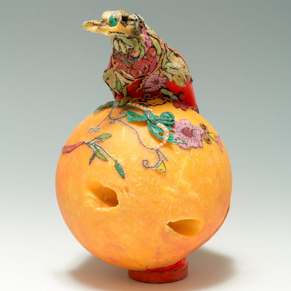 A waxy orange ball with a bird-like form sat on top of it. The bird is covered in floral embroidery scraps, which also partly cover the ball