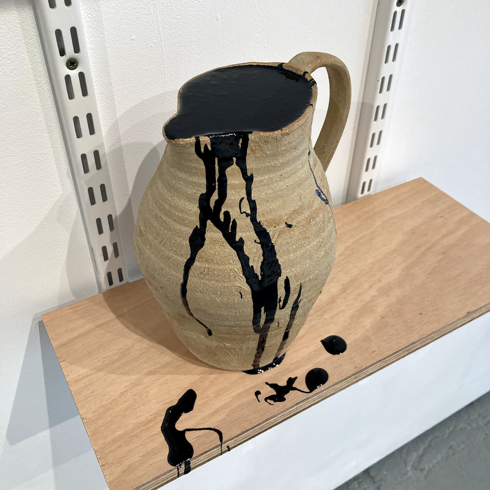 A ceramic jug made from rough, unglazed clay. A dark sticky liquid drips down the side and pools onto the self that its sitting on