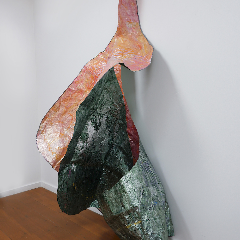 An abstract tubular sculpture made from paper. The outer surface is painted with green metallic paint and is torn to reveal the inner surface. which is metallic pink.