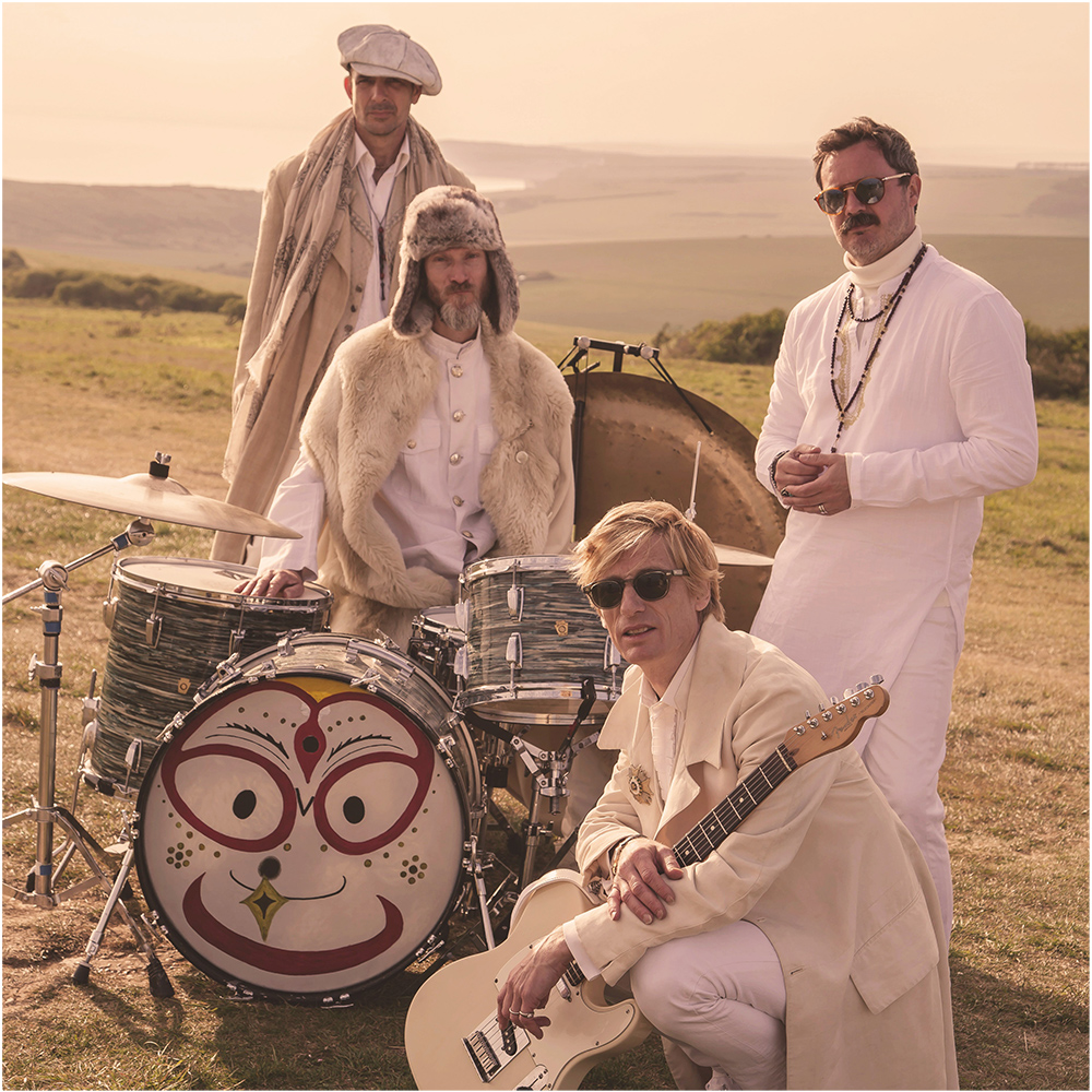Four men dressed in white clothes pose with their musical instruments (a drum, gong and electric guitar) in a field.