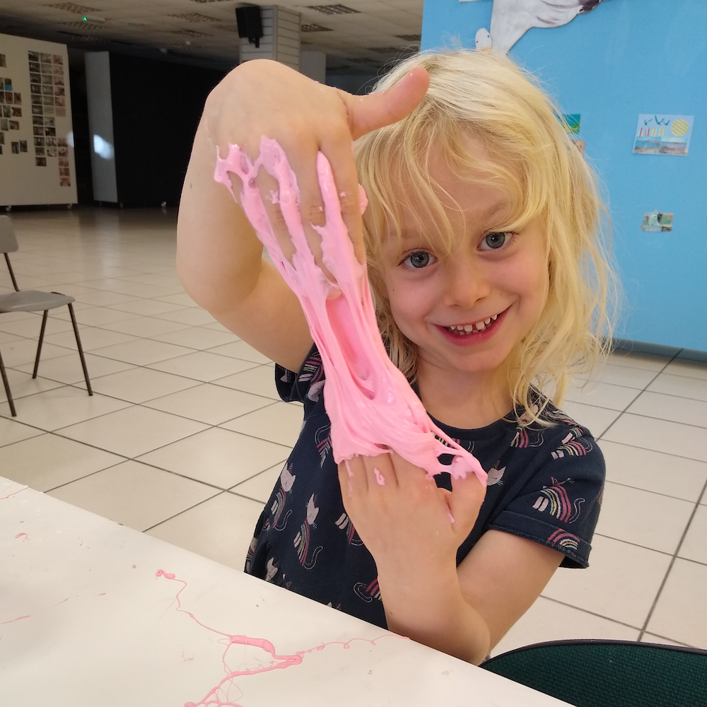 A young girl stretches a pink slime solution in her hands. She is sat at a table. There is a blue board behind her covered in a seaside themed decoration