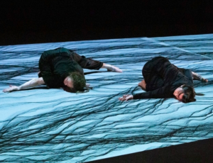 Two dancers lie on a screen on the floor which has wave animations on it
