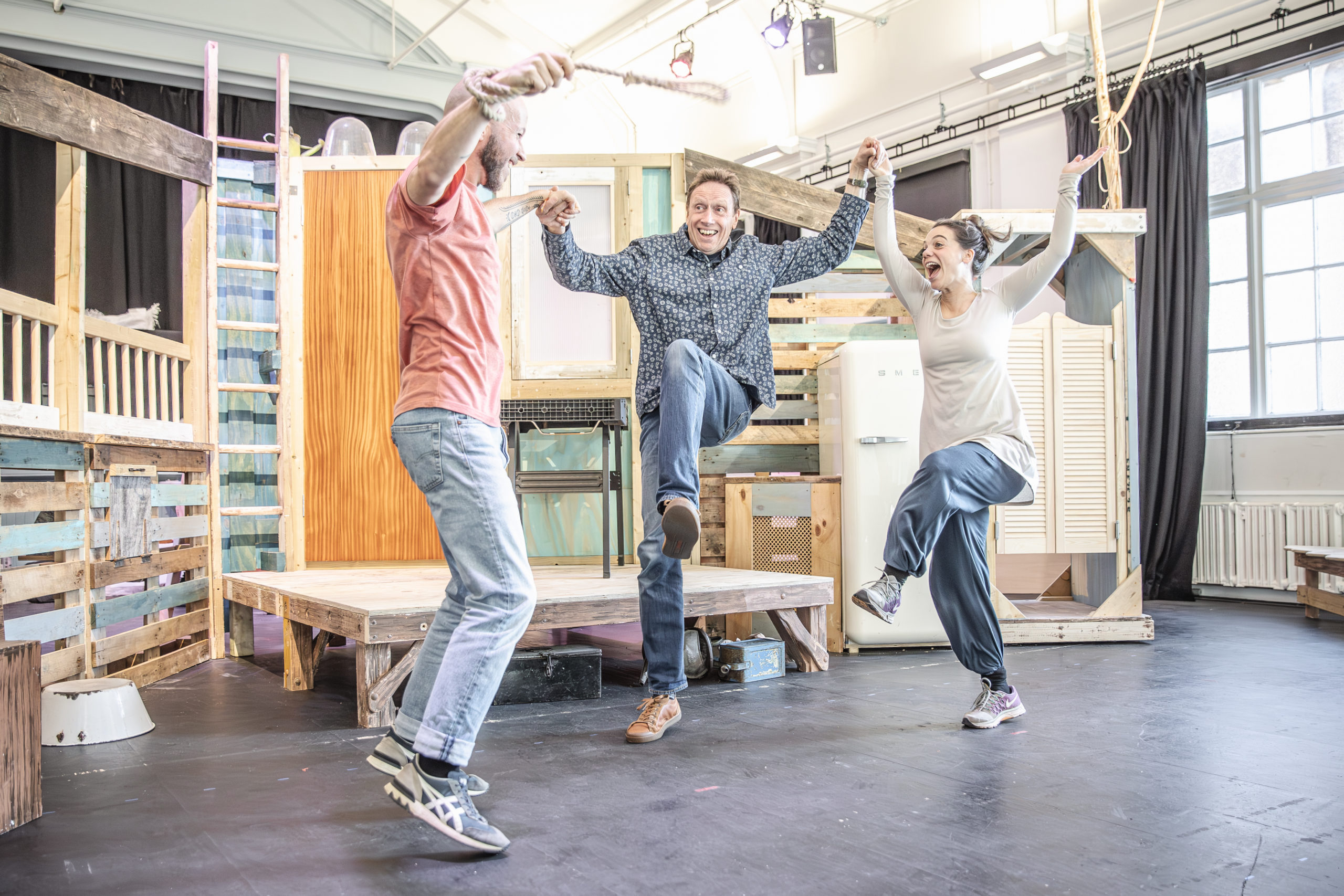 Three performers hold hands and jump in front of a set of a house made from recycled materials