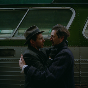 Two men in old fashioned clothing hug in front of an old train or bus carriage