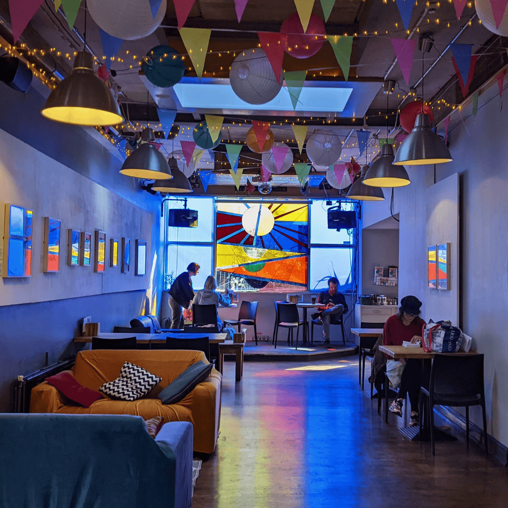 Exeter Phoenix Cafe Bar interior with colourful bunting and string lights on the ceiling. A large stained-glass window (by artist Terry Frost) casts a blue, red and yellow light into the room, while patrons sit on sofas and to small tables.
