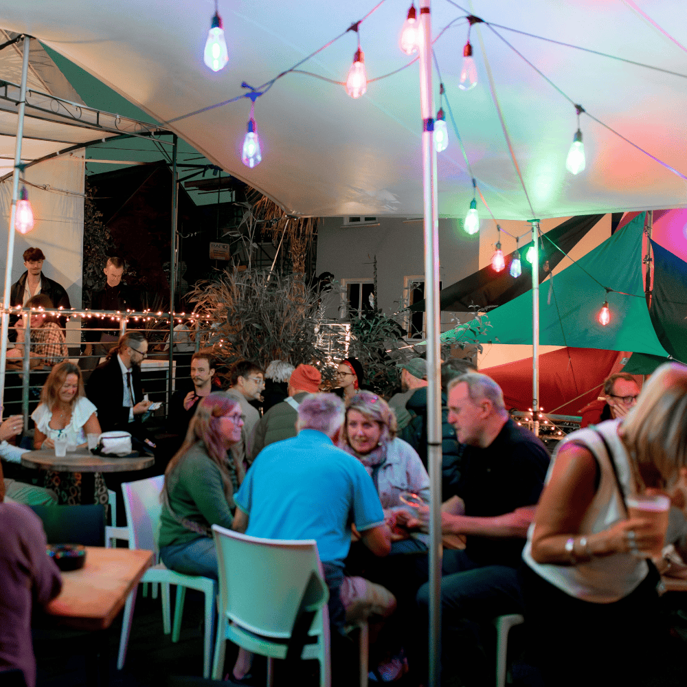 Outside on Exeter Phoenix's Cafe Bar Terrace, in the evening under a large white canopy with colourful string lights. The image features a crowd of adults seated around small tables and standing, holding drinks and having conversations.
