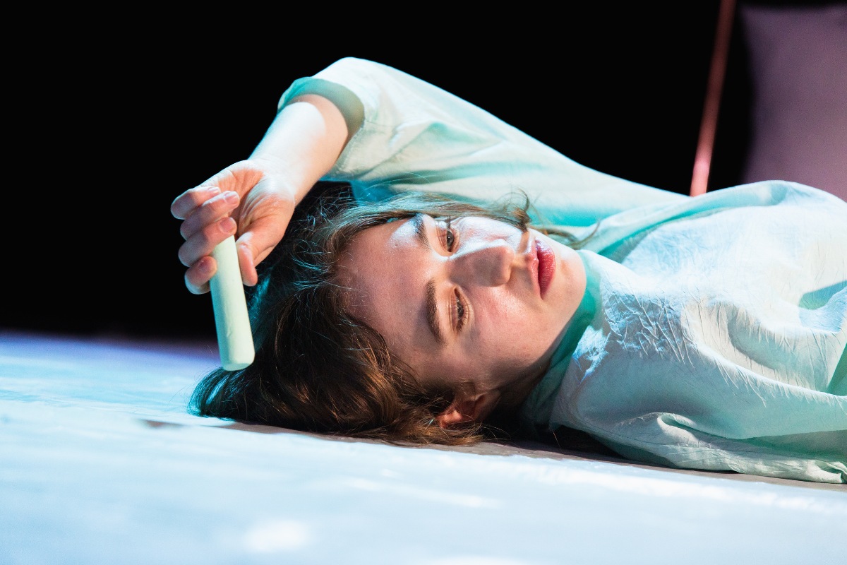 The image is a close up shot of the performer’s face against a dark background. The performer is white afab and has their eyes downcast. The performer is lying on the floor with their left arm reaching over the top of their head and their left hand holding a piece of light blue chalk. This image symbolises a moment in the show when the performer is drawing a chalk circle around themselves creating a physical outline of the body connected to how trauma is held in the body.