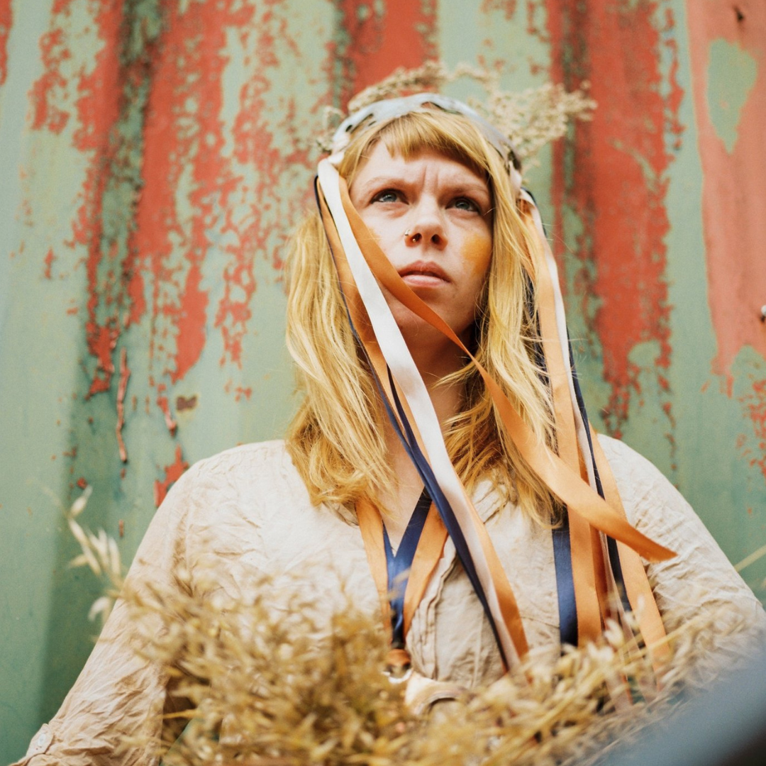Woman with ribbons all over, wheat in front and rusted metal behind them.