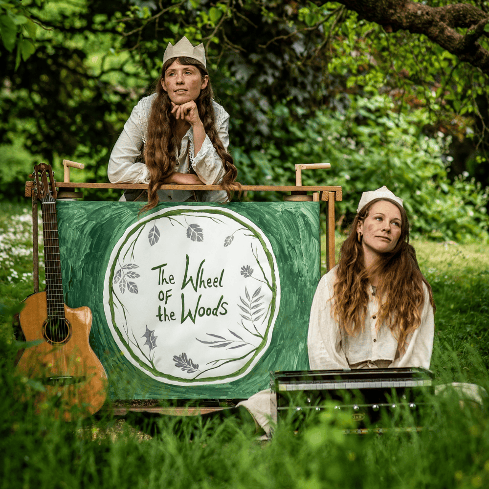 Two performers sat in some long grass with a guitar and accordian, and a crankie painting that says 'The Wheel of the Woods'
