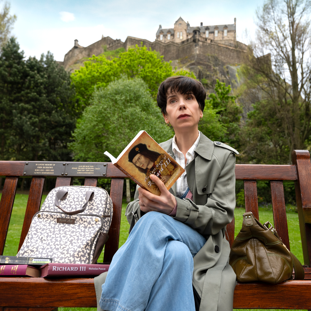 A woman with short hair is sat on a bench reading. Shes surrounded by Richard iii books and behind her is a castle and park