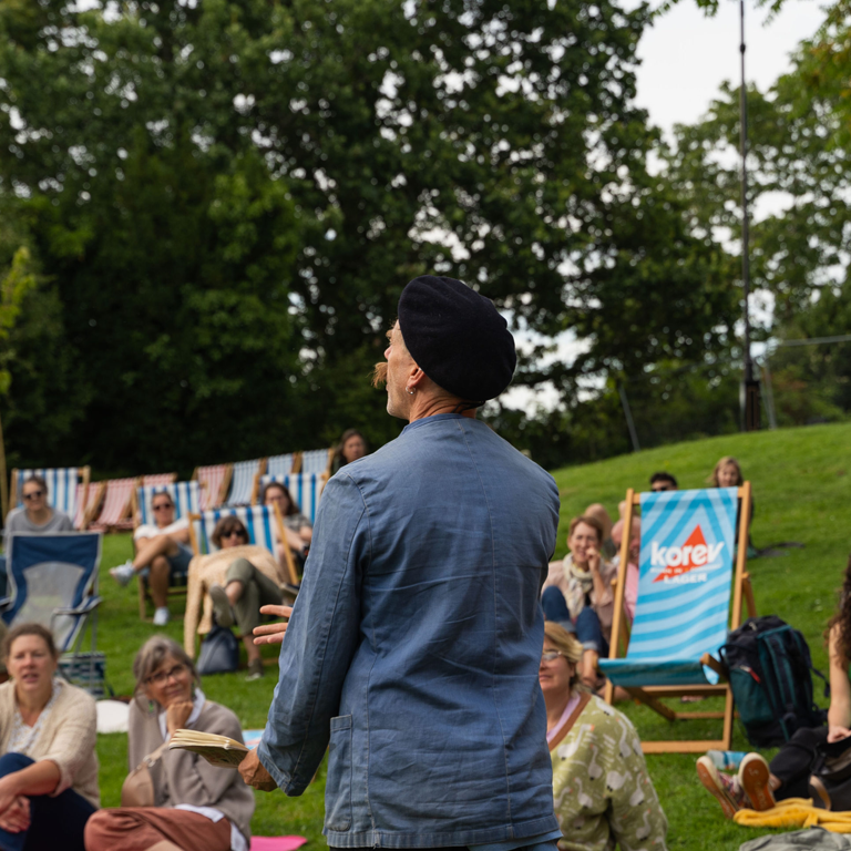 Someone performing at an open mic in rougemont gardens, with the audience sat on picnic blankets